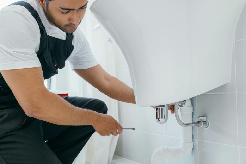 In a bathroom a technician repairs a water pipe under the sink. The skilled plumber focuses on